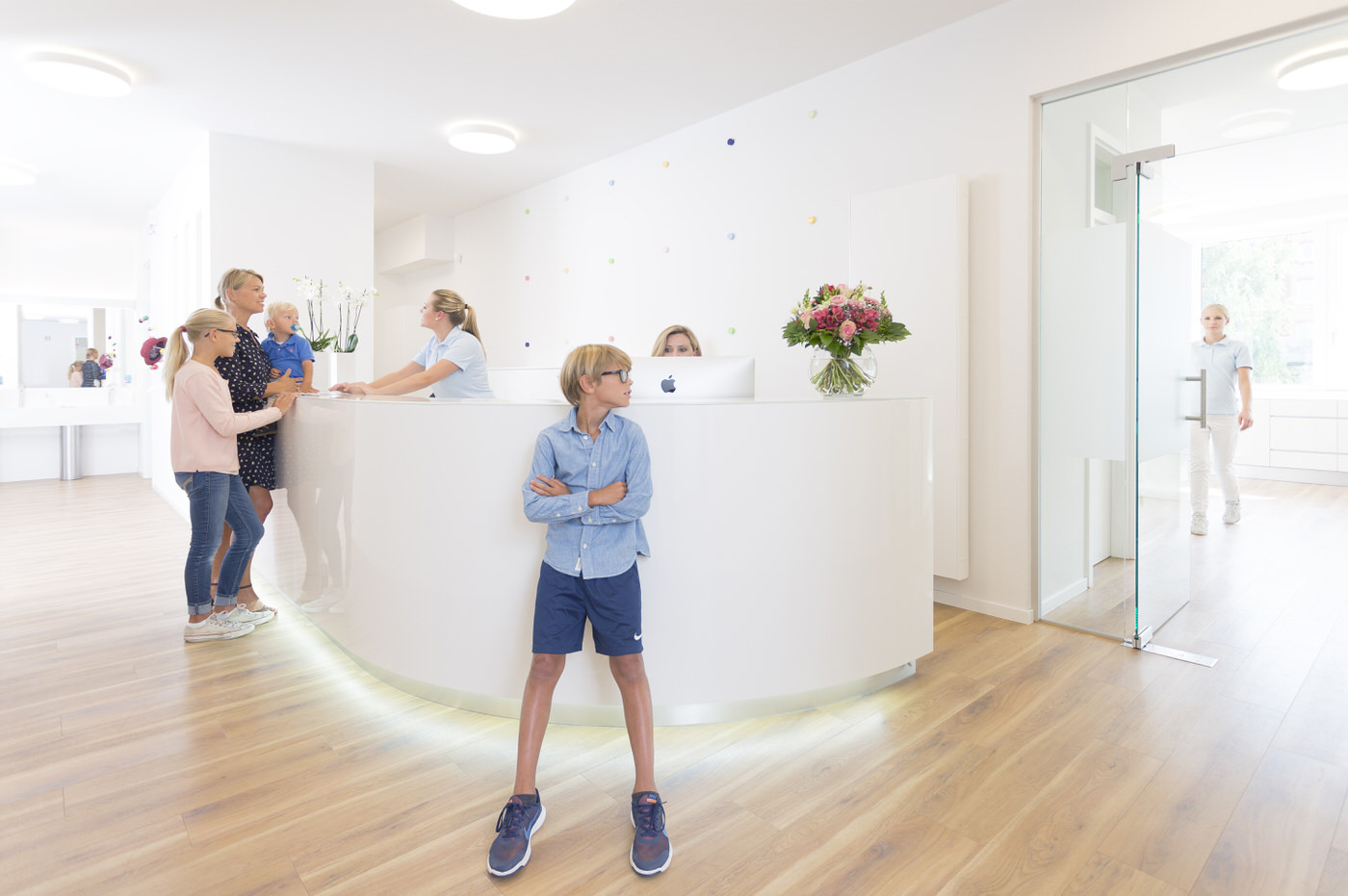 Helle, moderne Empfangssituation in einer Kinderarztpraxis. Eine Mutter mit drei Kindern steht an der runden, weißen Rezeption und spricht mit einer Mitarbeiterin. Ein Junge steht mit verschränkten Armen im Vordergrund. Eine weitere Mitarbeiterin schaut hinter einem Computer hervor, während im Hintergrund eine dritte Frau durch eine Glastür kommt. Die Atmosphäre wirkt freundlich und einladend, mit hellem Holzfußboden, dezenten Farben und frischen Blumen.