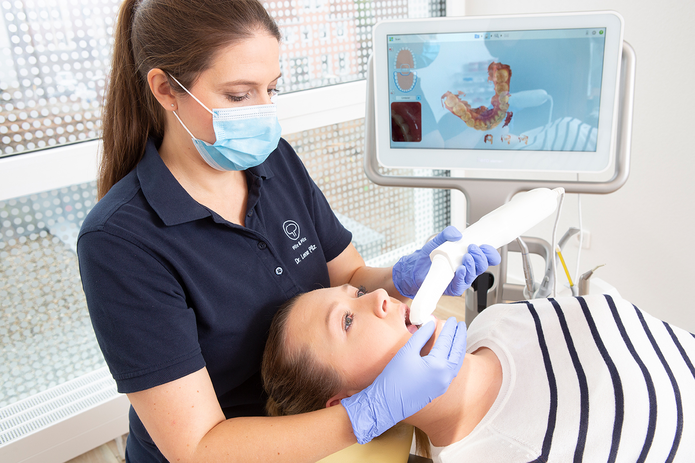 Behandlungszimmer in einer modernen Zahnarztpraxis mit heller, minimalistischer Einrichtung. Eine junge Patientin sitzt auf dem Behandlungsstuhl, während die Zahnärztin mit Schutzmaske konzentriert mit einem oralen 3D-Scannerarbeitet. Im Hintergrund sieht man das teilweise Erstellte Bild des 3D-gescannten Gebisses der Patientin.