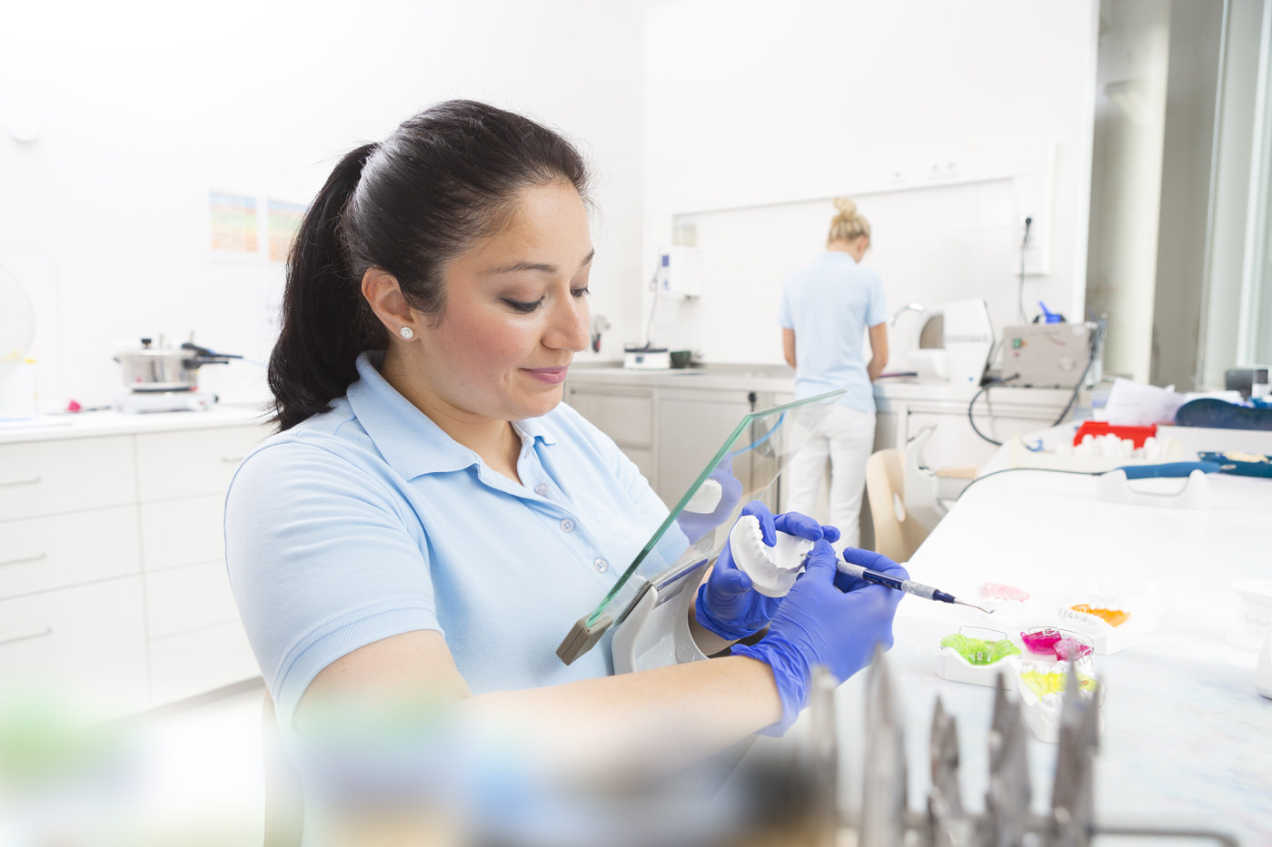 Zahntechnikerin bei der präzisen Farbgestaltung eines Zahnmodells im Praxislabor. Mit blauen Handschuhen und konzentriertem Blick trägt sie Farbe auf ein dentaltechnisches Modell auf – moderne Handarbeit trifft auf höchste Präzision in der Zahntechnik.