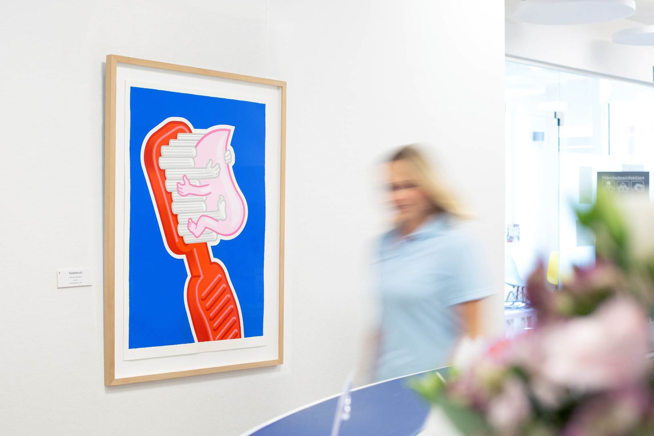 Kunst im Praxisflur: Ein farbenfrohes Bild mit roter Zahnbürste und rosa Figur vor kräftig blauem Hintergrund hängt an der Wand einer modernen Zahnarztpraxis. Eine Mitarbeiterin geht unscharf durchs Bild, im Vordergrund sind Blumen angedeutet.