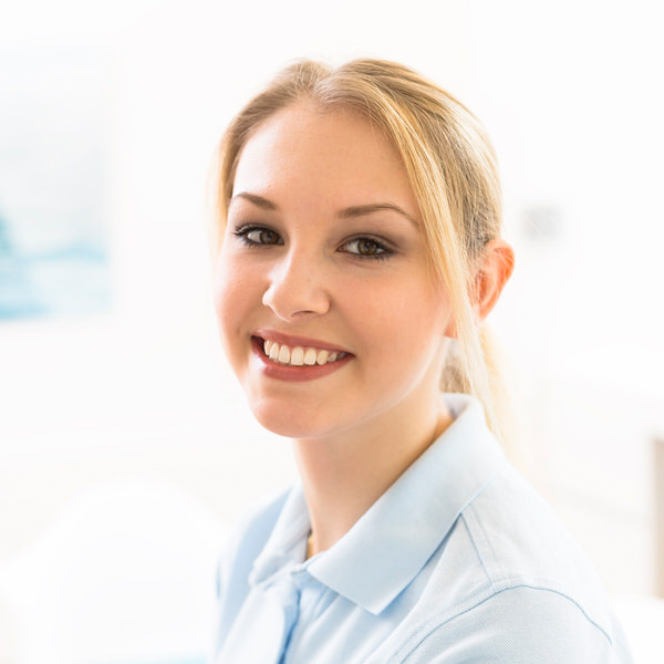 Porträt von Jennifer Lux vor einem unscharfen Hintergrund. Sie trägt ein hellblaues Polo-Shirt und lächelt freundlich in die Kamera