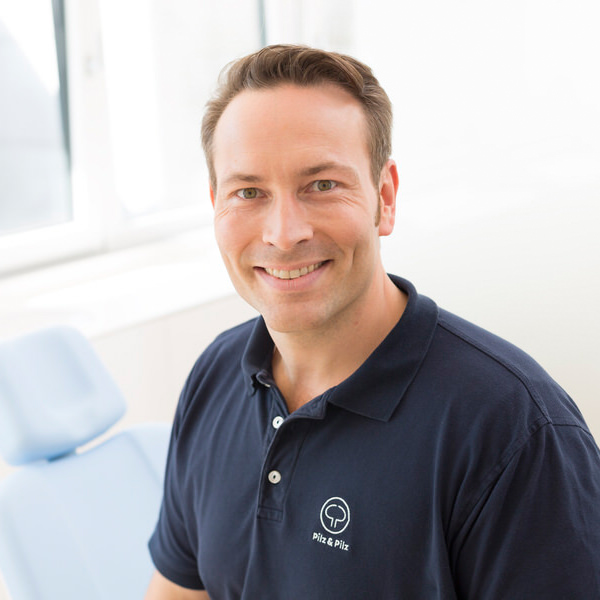 Porträt von Dr. Peter Pilz vor einem unscharfen Hintergrund. Er trägt ein dunkelblaues Polo-Shirt mit weiß aufgesticktem Pilz-&-Pilz-Logo und lächelt freundlich in die Kamera