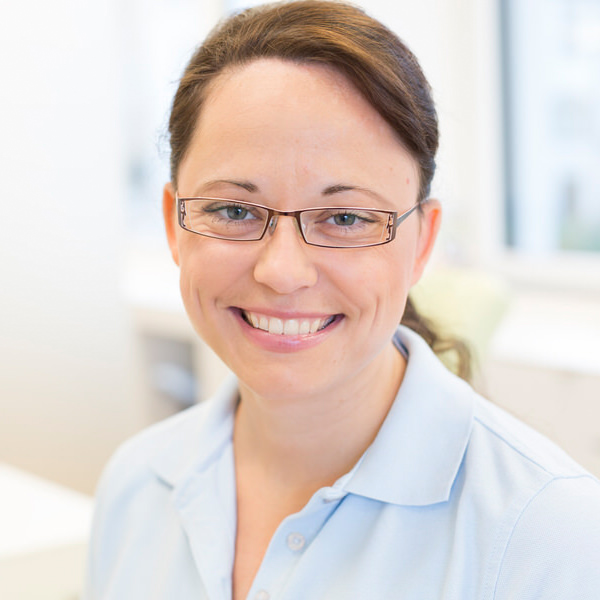 Porträt von Regina Merk vor einem unscharfen Hintergrund. Sie trägt ein hellblaues Polo-Shirt und lächelt freundlich in die Kamera