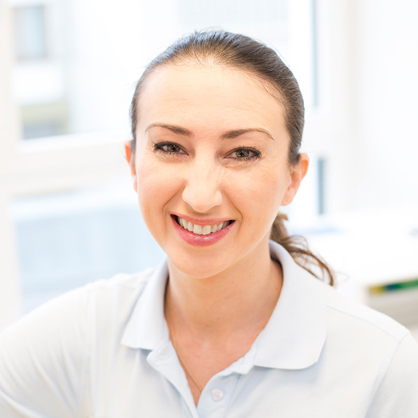 Porträt von Tatjana Hart vor einem unscharfen Hintergrund. Sie trägt ein hellblaues Polo-Shirt und lächelt freundlich in die Kamera